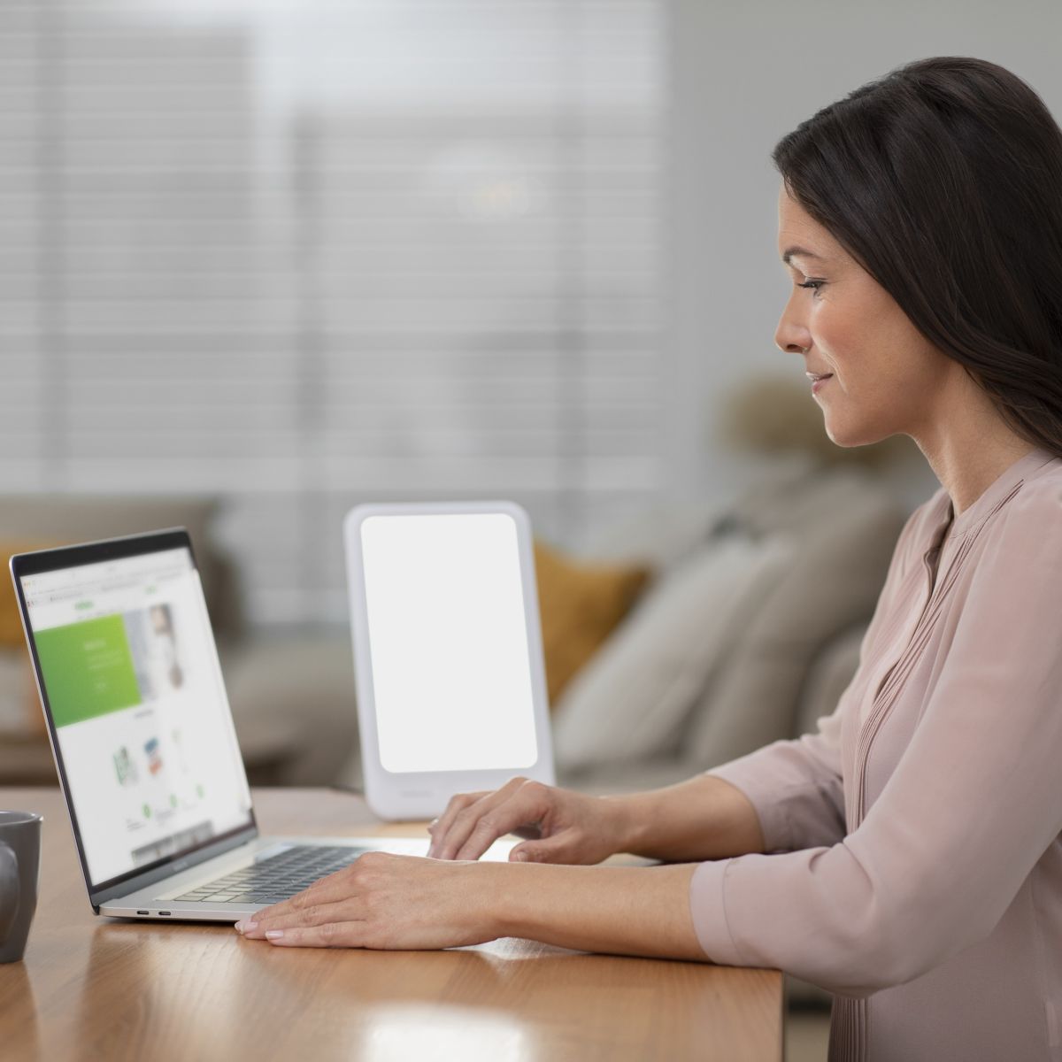 Eine Frau sitzt am Tisch und arbeitet am Laptop, während eine eingeschaltete Tageslichtlampe für helles, gleichmäßiges Licht sorgt.